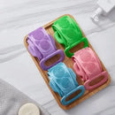 Four colorful silicone scrubbers on a wooden tray with a white towel in the background.