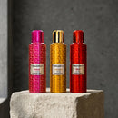 Three Blackberry essential oil spray bottles in pink, gold, and red, standing on a stone pedestal against a dark gray background.